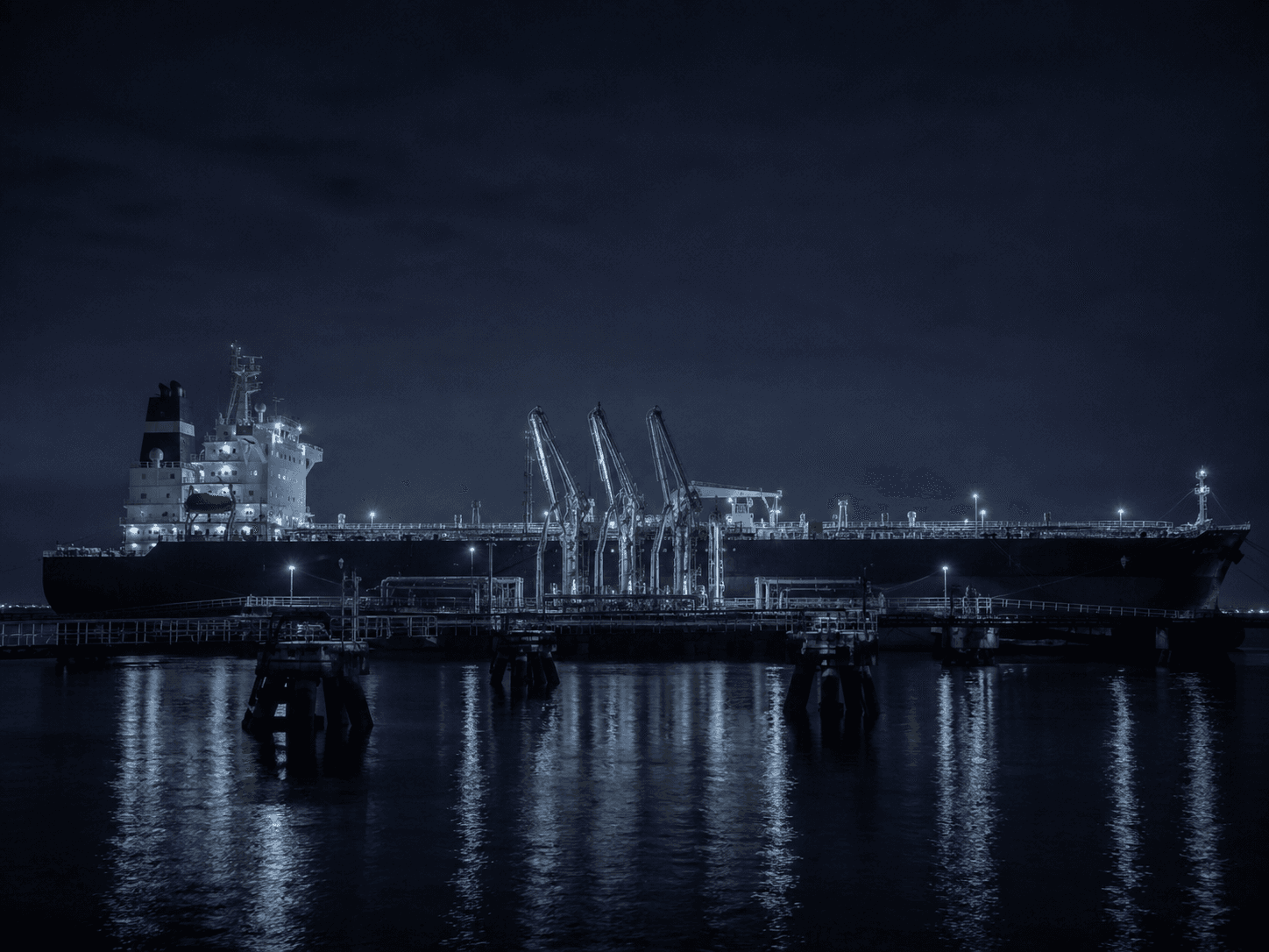A crude tanker berthed at a marine loading jetty at night, shore loading arms connected to the midship manifold.